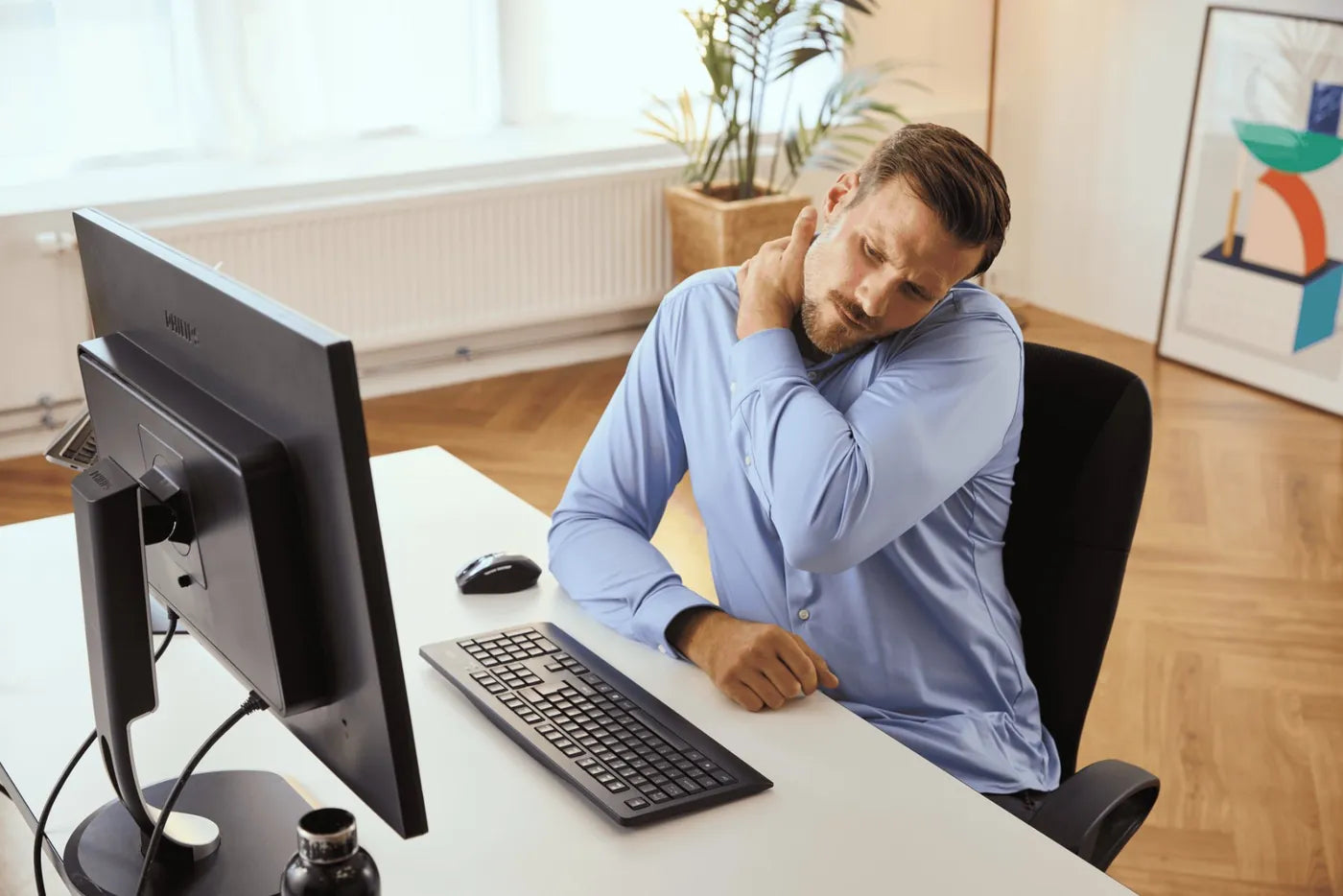 A man at his desk rubbing his shoulder. A standard mouse sits off to the right of his keyboard.